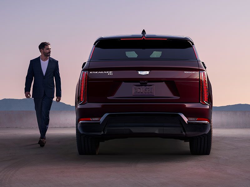 A Man Walking Beside the Rear End of a 2026 ESCALADE IQ Parked in the Desert During Dusk
