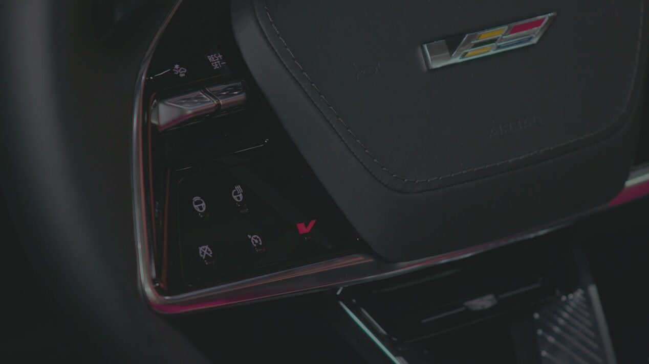A Close-Up View Of A Cadillac OPTIQ Steering Wheel Showing Illuminated Touch-Sensitive Control Icons Beside the Lower Spoke.