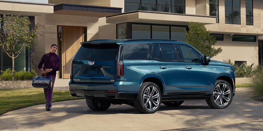 Three Quarters View of a Blue Cadillac SUV Parked in Front of a House While a Man Walks to the Trunk with a Bag