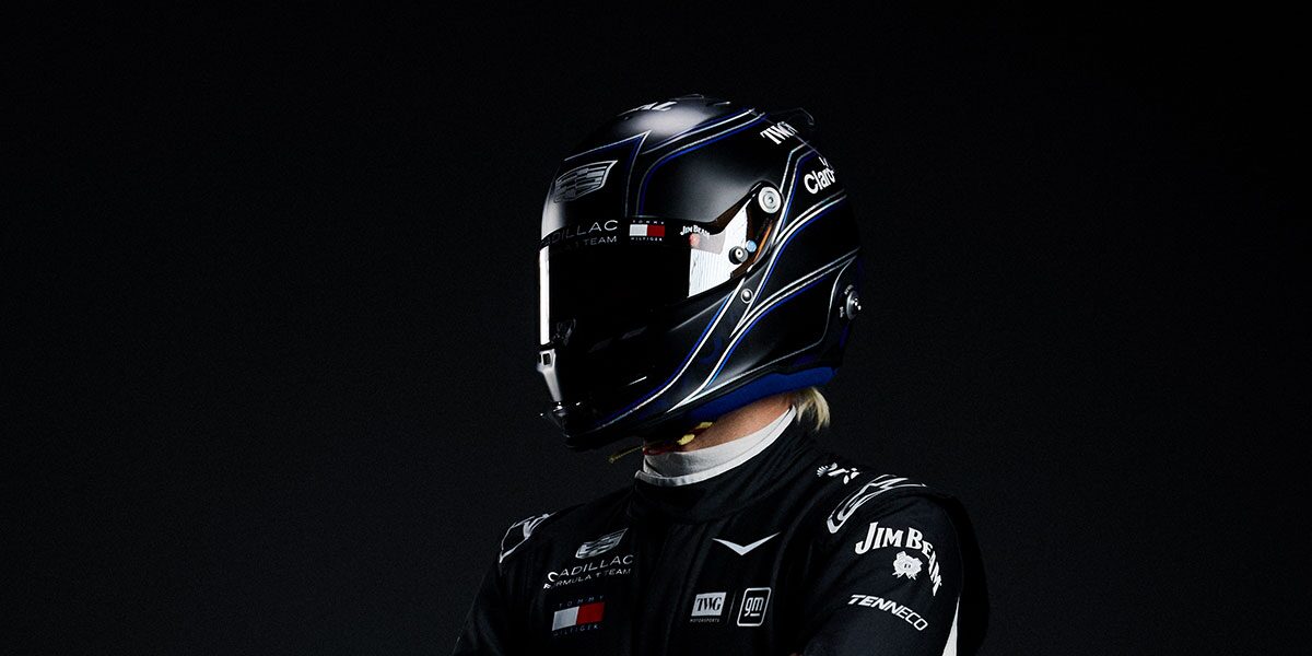 A Driver Wearing A Glossy Black Racing Helmet And A Black Cadillac Race Suit Stands Against A Dark Studio Background.