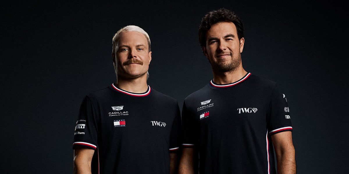 Two Team Members In Black Cadillac Racing Suits Stand Shoulder To Shoulder Against A Dark Studio Background.