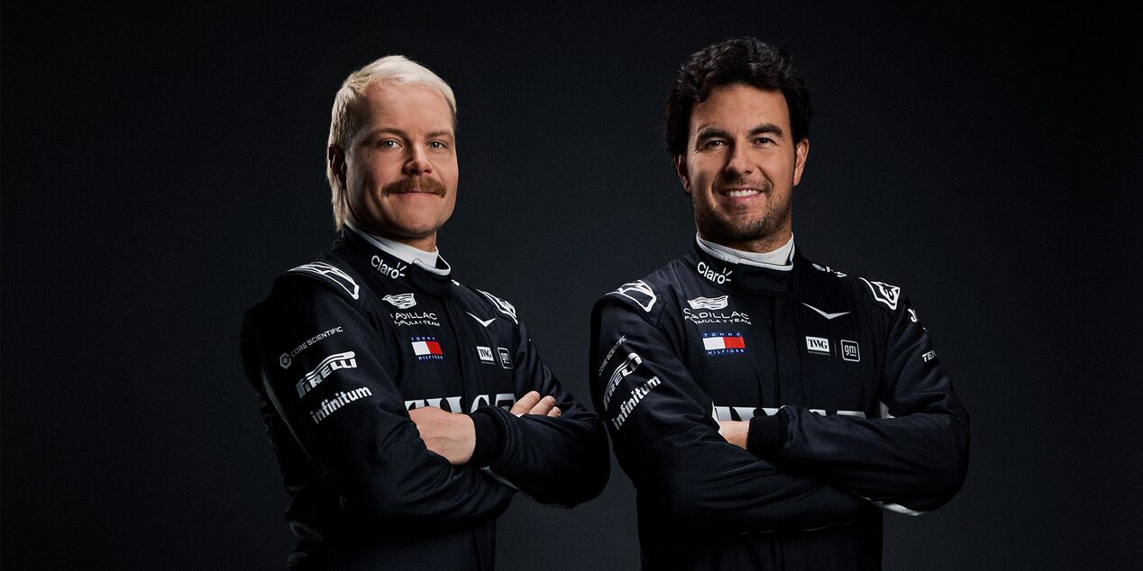 Two Team Members In Black Cadillac Racing Suits Stand Shoulder To Shoulder Against A Dark Studio Background.