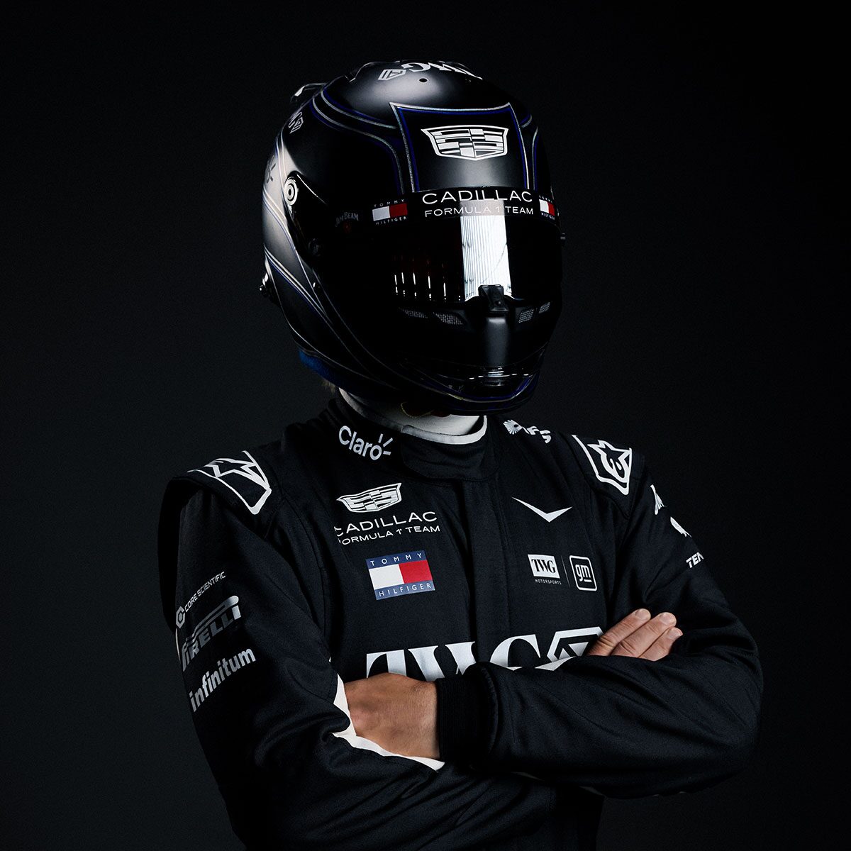 A Driver In A Black Cadillac Race Suit And Glossy Black Helmet Stands With Arms Crossed Against A Dark Studio Backdrop.
