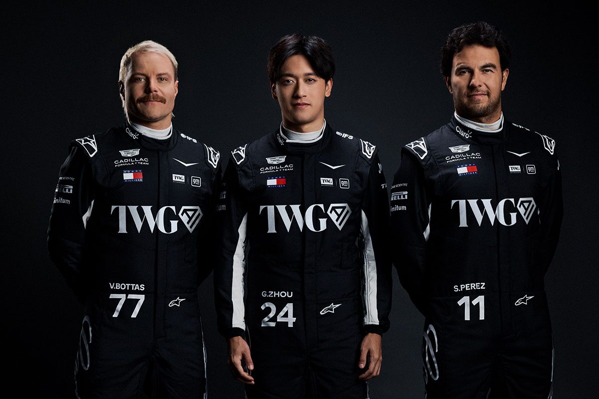 Three Team Members Wearing Matching Black Racing Suits With Cadillac And Sponsor Logos Stand Side By Side Against A Dark Studio Background.
