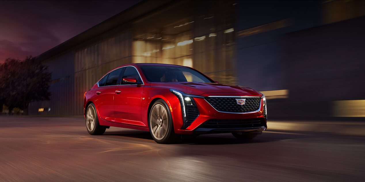 Three-quarters Front View of a Red 2025 Cadillac CT5 Driving Past an Industrial Building