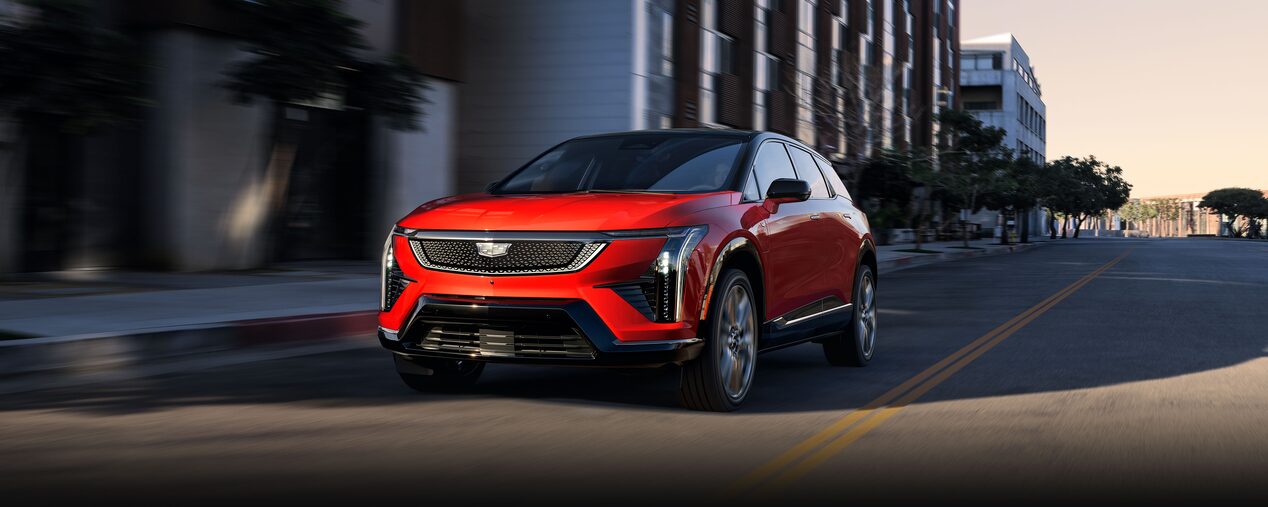 A Red 2026 Cadillac OPTIQ Driving Down a City Street with Buildings in the Background