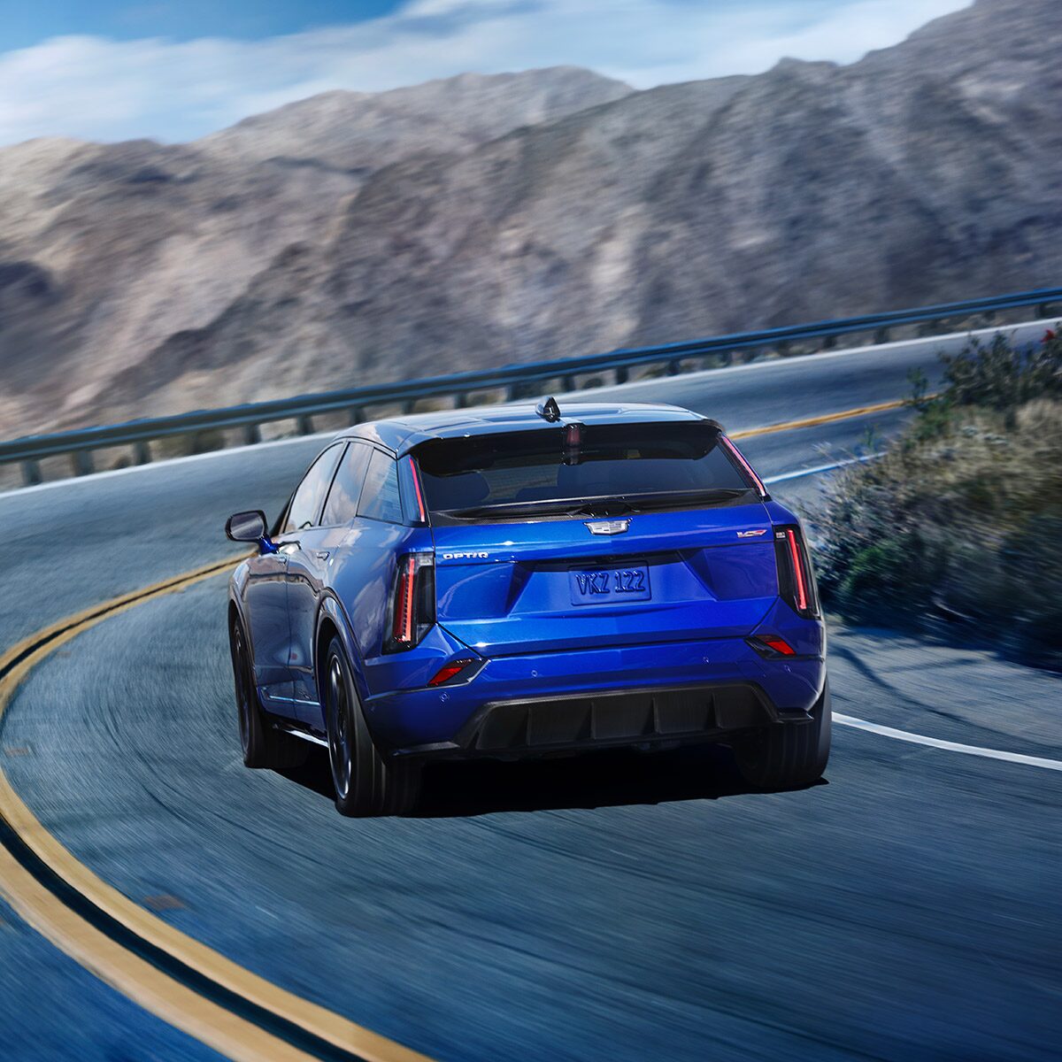 Rear View of a Blue 2026 OPTIQ-V Driving on a Curve of a Mountain Row