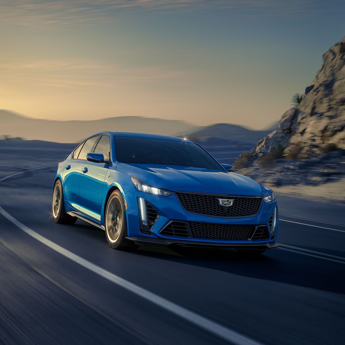A Blue Cadillac V-Series Car Driving Down a Highway with Mountains in the Background.