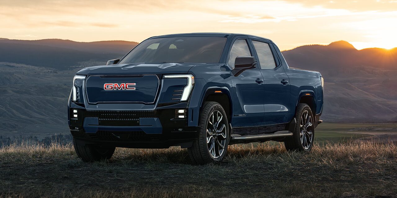 Front Three-Quarters View of the 2026 GMC Sierra EV Parked on a Grassy Hill at Sunrises Over the Mountains