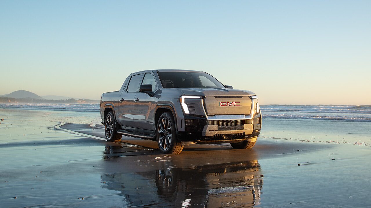 A Gray GMC Sierra EV Pickup Truck Parked on a Wet Sandy Beach at Sunset with the Ocean in the Background