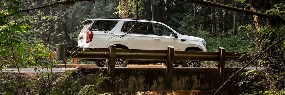 Side View of a GMC White SUV Crossing a Small Bridge Surrounded by Trees