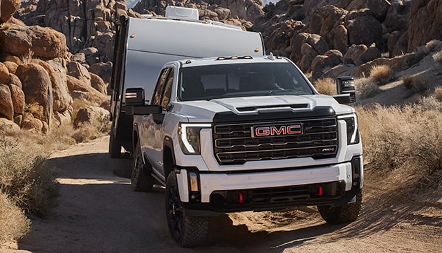 A White GMC Sierra HD AT4X Truck Towing a Silver Travel Trailer on a Dirt Road Through a Rocky Desert