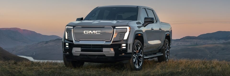 The GMC Sierra EV pickup truck parked in a green valley at sunset with a beautiful mountain view in the distance.