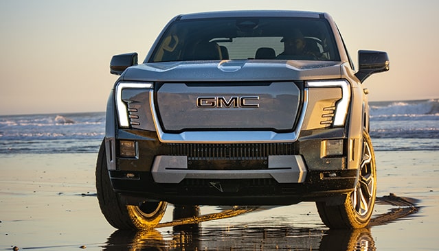 The Front View of a Gray GMC Sierra EV Truck Parked on a Sandy Beach at Sunset, with Ocean Waves in the Background