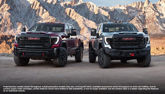 A Red GMC Sierra HD AT4X and a Gray GMC Sierra HD AT4X AEV Edition Pickup Truck Parked on a Dirt Road with Snowy Mountains in the Background
