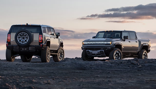 A Beige GMC HUMMER EV SUV and a Gray GMC HUMMER EV Pickup Parked on a Dirt Hill at Sunset