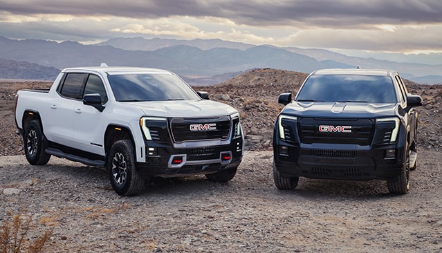 A White GMC Canyon AT4X and a Dark GMC Canyon AT4X AEV Edition Pickup Truck Parked on a Dirt Road in a Desert Landscape