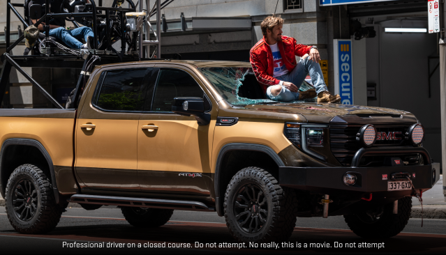 Ryan Gosling in a Red Jacket Sitting on the Hood of a Brown GMC Sierra Pickup Truck with a Broken Windshield, from the Movie Fall Guy