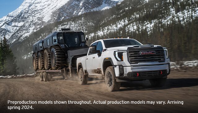 A White GMC Sierra HD Truck Towing a Large Multi-Wheeled Off-Road Vehicle on a Dirt Road in a Snowy Mountain Landscape