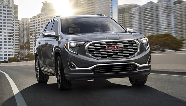 A Gray GMC Terrain SUV Driving on a Multi-Lane Highway With City Buildings in the Background