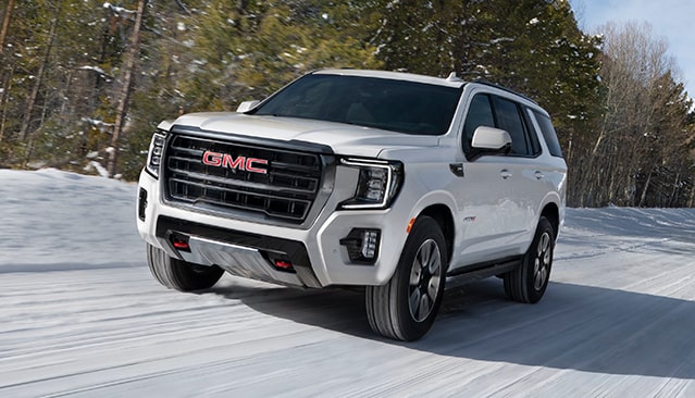 A White GMC Yukon AT4 SUV Driving on a Snowy Road Through a Forest of Bare Trees and Evergreen Pines