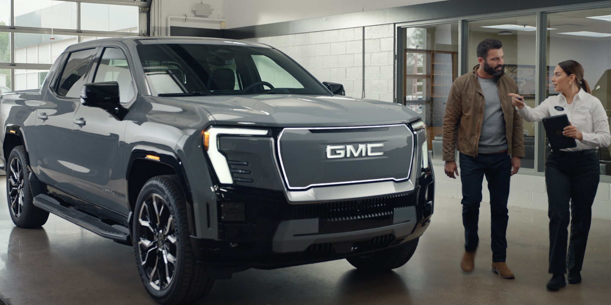 A Silver GMC Sierra EV Displayed in a Dealership Building with a Salesperson Showing a Customer the Details.