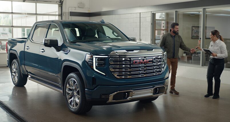 A Silver GMC Sierra EV Displayed in a Dealership Building with a Salesperson Showing a Customer the Details.
