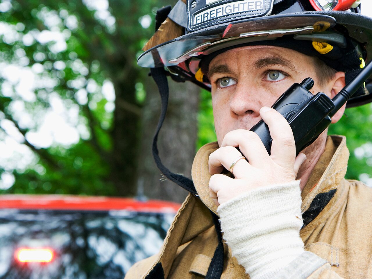 A Firefighter Wearing a Helmet and a Tan Jacket Is Holding a Two-Way Radio to His Ear In Front of a Red Emergency Vehicle