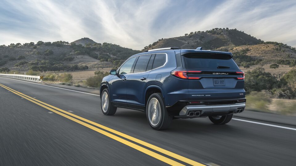 Rear View of the GMC Acadia Denali Driving Down the Open Road with Blue Skies in the Distance
