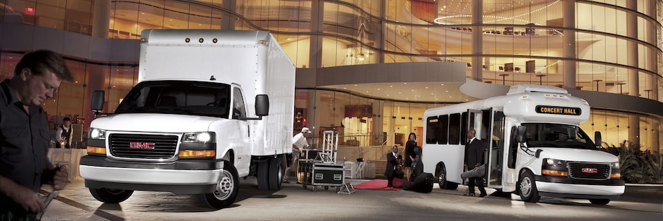 Two 2025 GMC Savana Cutaway Vans Parked Outside of a Glass Building at Night