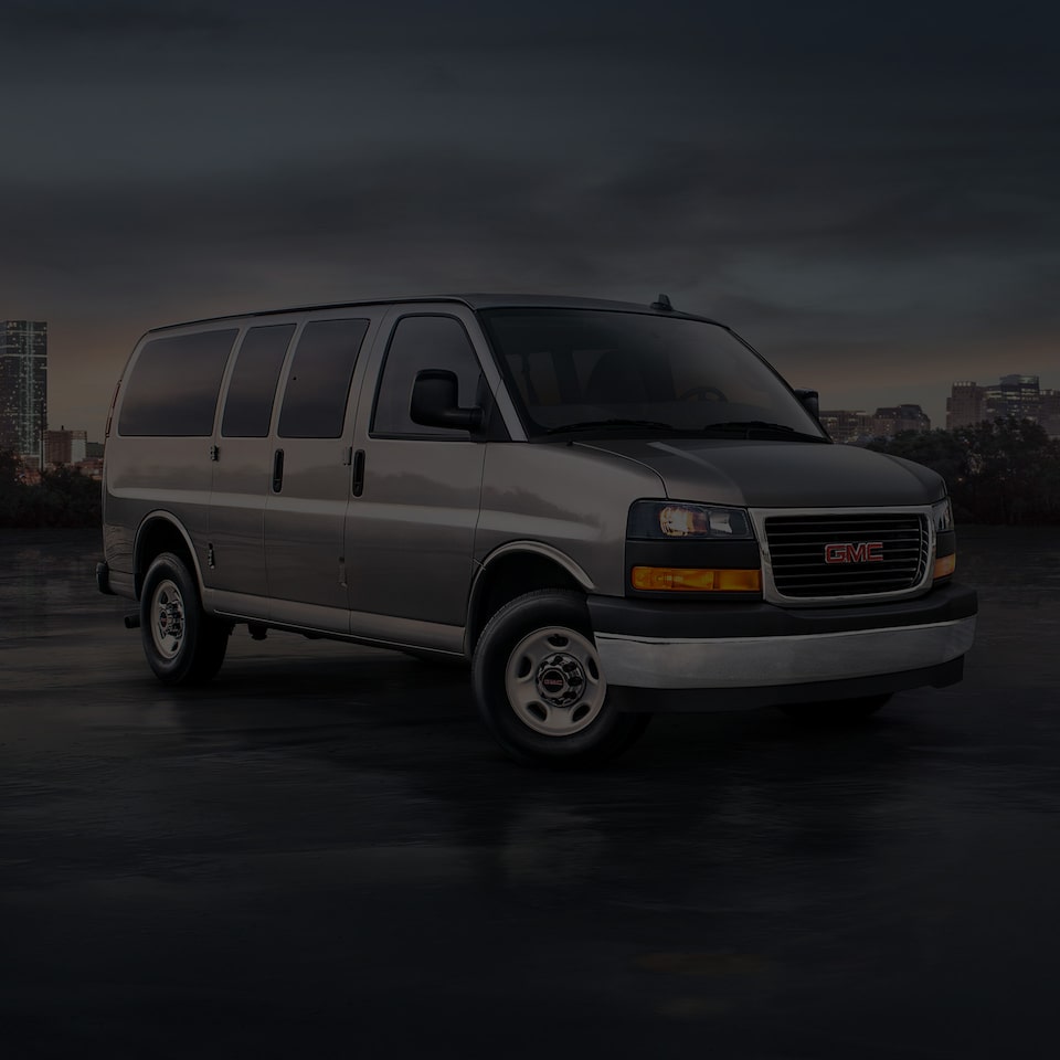 Front Three-Quarters View of a 2025 GMC Savana Passenger Van with a City in the Background