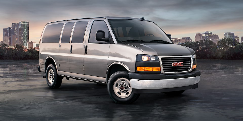 Front Three-Quarters View of a 2025 GMC Savana Passenger Van with a City in the Background