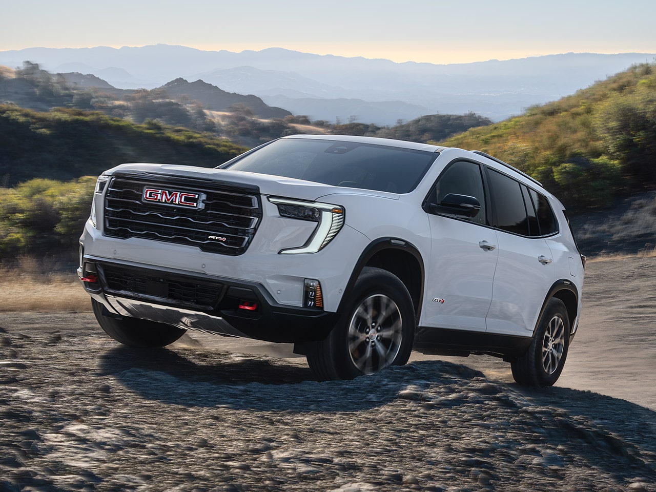Exterior View of the 2026 GMC Acadia AT4 Driving Over Rough Terrain with a Beautiful Mountain View in the Distance