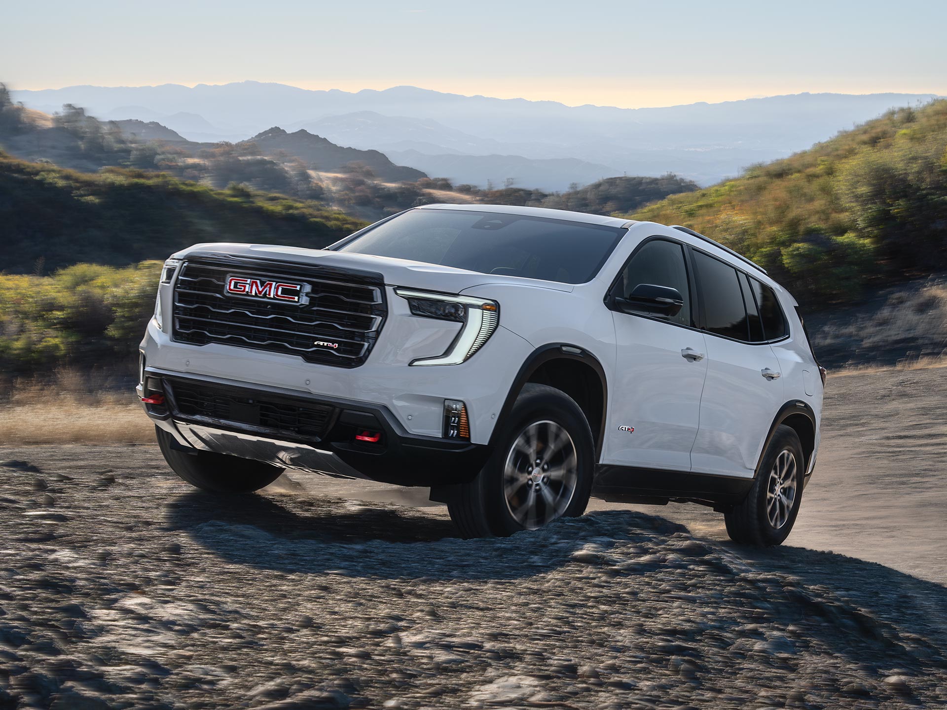 Exterior View of the 2026 GMC Acadia AT4 Driving Over Rough Terrain with a Beautiful Mountain View in the Distance