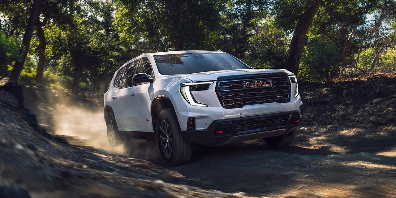 Exterior View of the 2026 GMC Acadia Mid-Size SUV Driving on a Dirt Path Surrounded by Trees