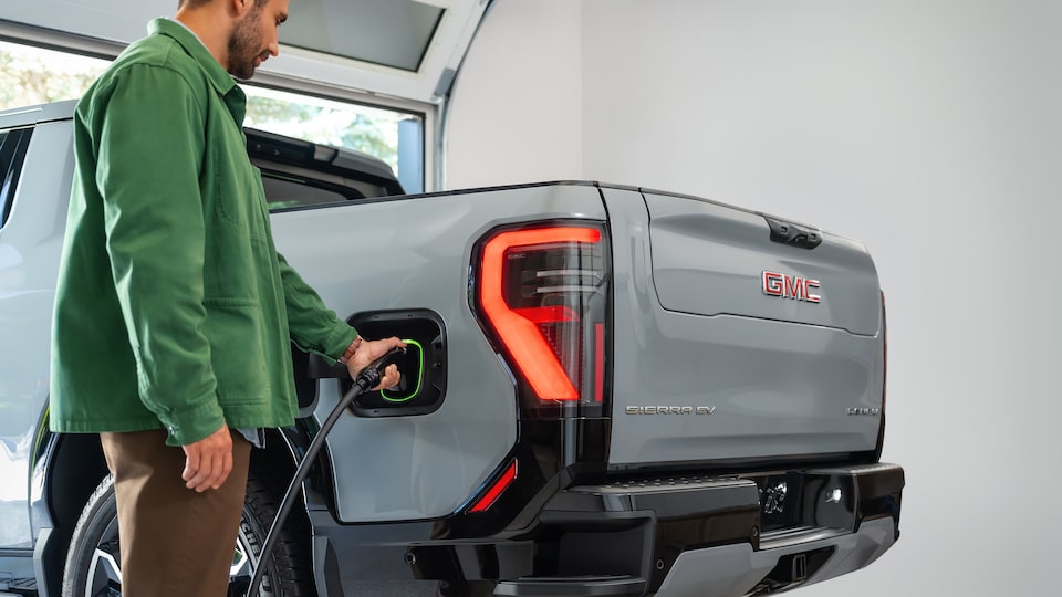A Man Plugging a Charging Cable into the Side of a Gray GMC Sierra EV Pickup Truck with an Illuminated Taillight and GMC Logo